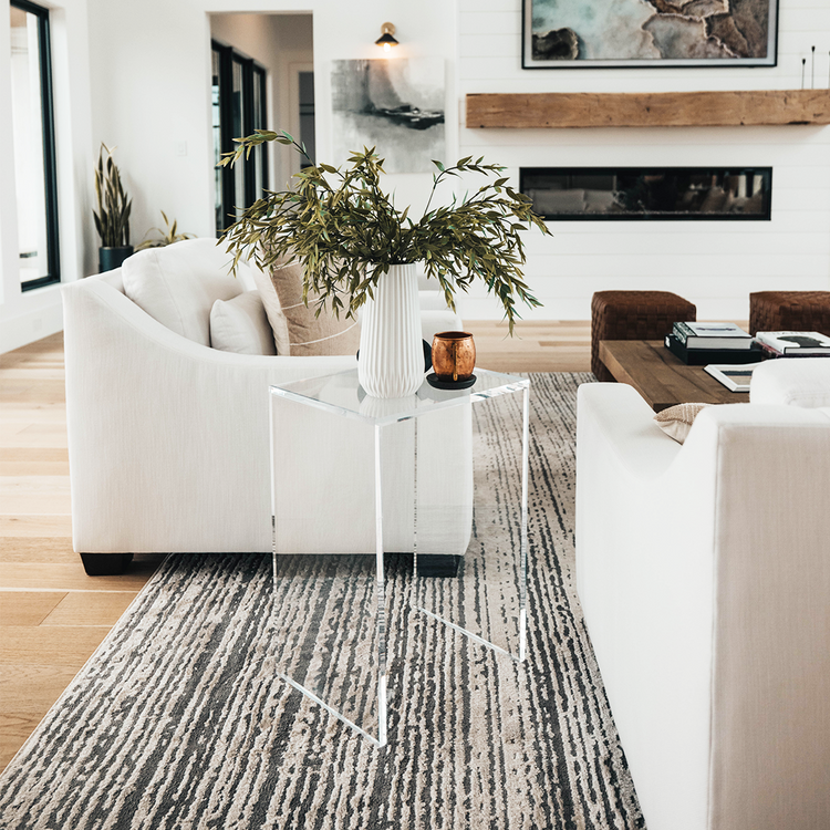 A vertical rectangular shaped acrylic side table holding a plant and a coffee cup placed on a rug between a white couch and a white sofa in a modern living room.