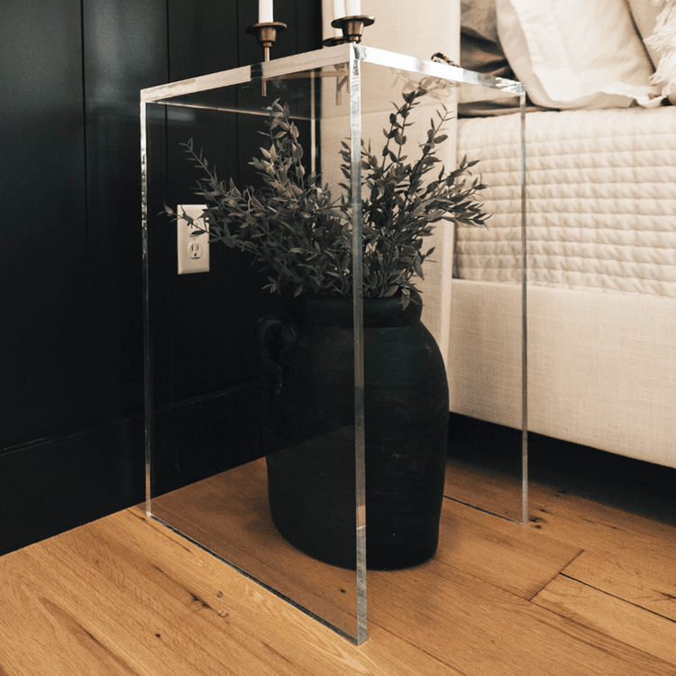 A vertical rectangular shaped acrylic side table holding candles with a vase of flowers slid underneath placed beside a bed.