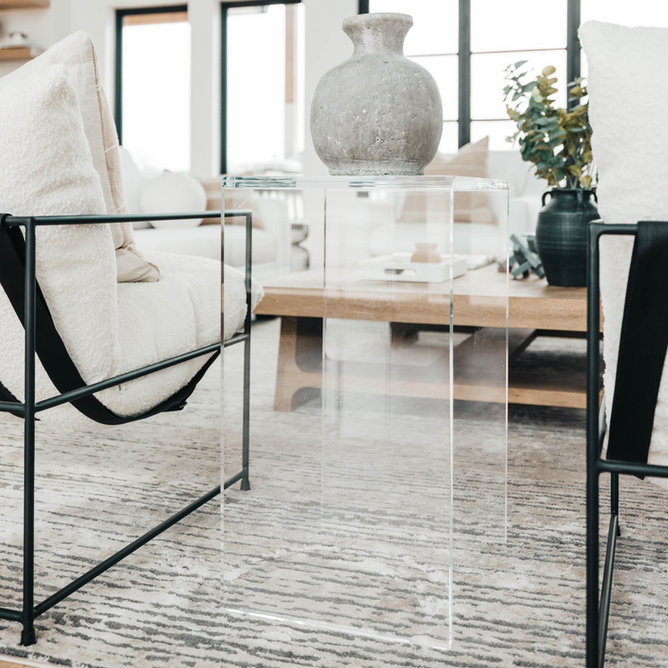 A vertical rectangular shaped acrylic side table holding a ceramic vase placed on a rug between two chairs in a modern living room.