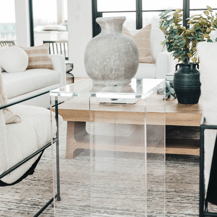 A vertical rectangular shaped acrylic side table holding a ceramic vase placed on a rug between two chairs in a modern living room.