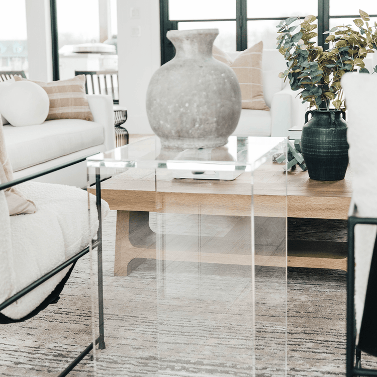 A vertical rectangular shaped acrylic side table holding a ceramic vase placed on a rug between two chairs in a modern living room.