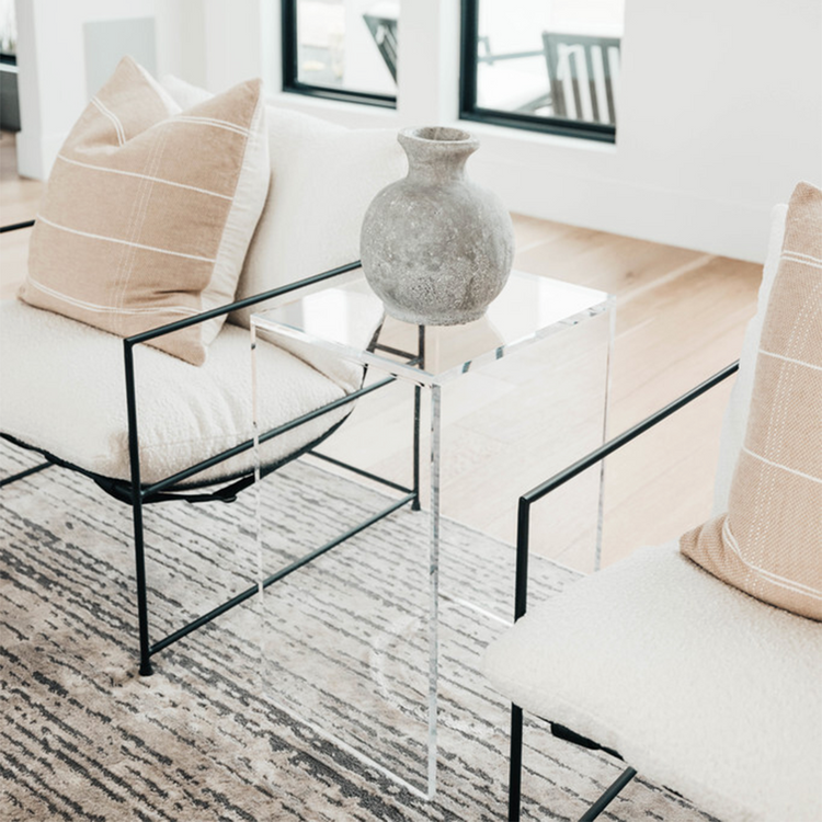 A vertical rectangular shaped acrylic side table holding a ceramic vase placed on a rug between two chairs in a modern living room.