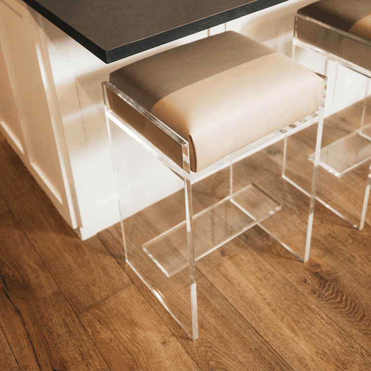 A close up of two stools out of a set of four clear acrylic modern bar stools with beige upholstered seats and clear acrylic legs, positioned in a kitchen setting.