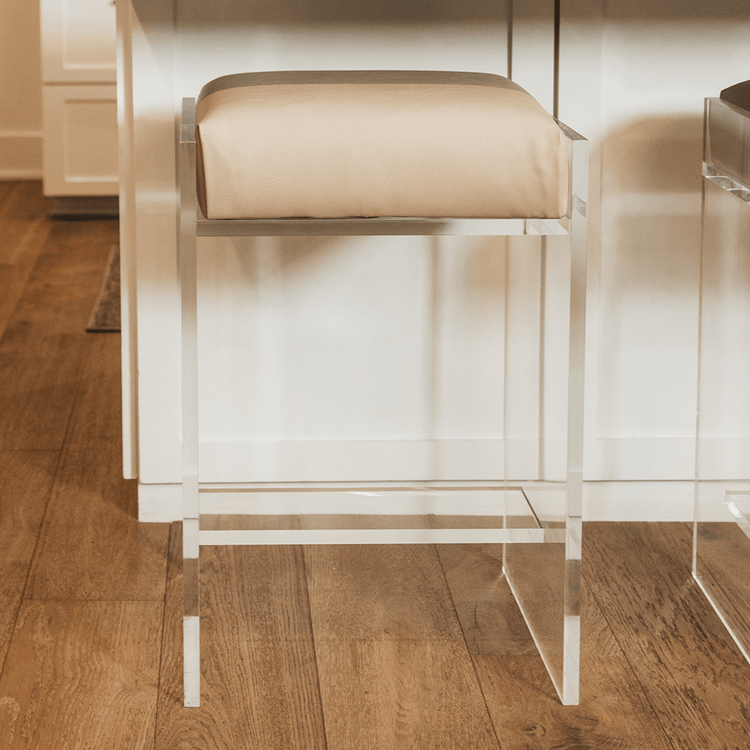A close up of one stool out of a set of four clear acrylic modern bar stools with beige upholstered seats and clear acrylic legs, positioned in a kitchen setting.