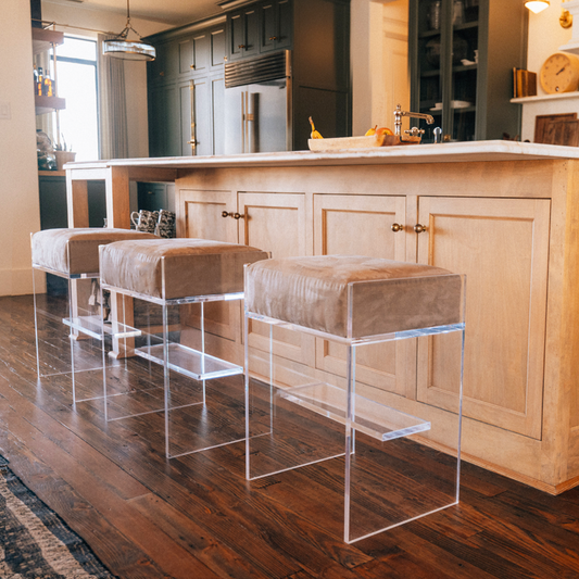 Three clear acrylic bar stools with beige velvet cushions at an island in a kitchen.