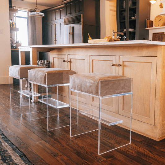 Three clear acrylic bar stools with beige velvet cushions at an island in a kitchen.
