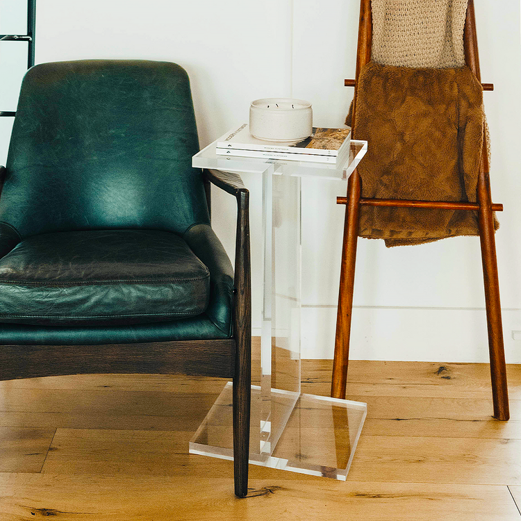 A clear acrylic side table shaped as the letter "I", positioned on a floor between a leather armchair and blanket stand displaying a candle and 2 books.