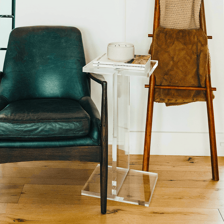 A clear acrylic side table shaped as the letter "I", positioned on a floor between a leather armchair and blanket stand displaying a candle and 2 books.
