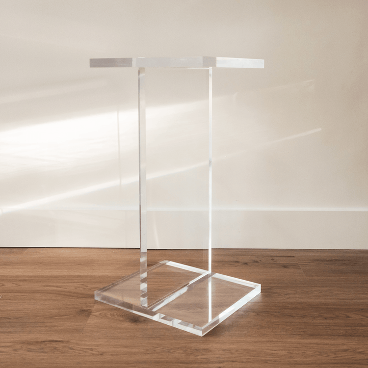 A catalogue view of a clear acrylic side table shaped as the letter "I", positioned on a hardwood floor.