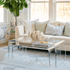 A clear acrylic lucite nesting coffee table set placed in a living room setting in front of a white sofa displaying decorative items on top and books on the smaller nesting table underneath.
