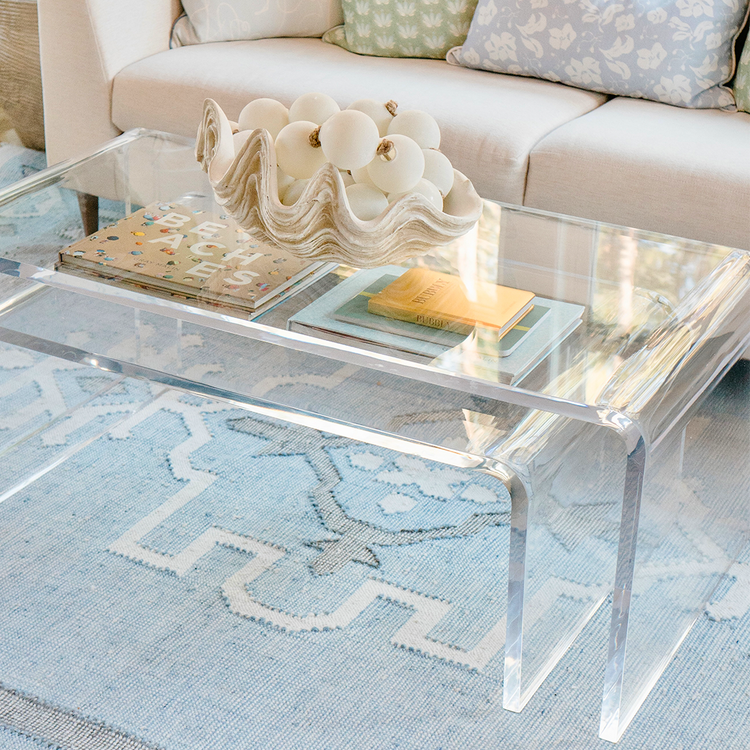 A clear acrylic lucite nesting coffee table set placed in a living room setting in front of a white sofa displaying decorative items on top and books on the smaller nesting table underneath.