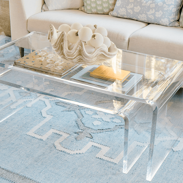 A clear acrylic lucite nesting coffee table set placed in a living room setting in front of a white sofa displaying decorative items on top and books on the smaller nesting table underneath.