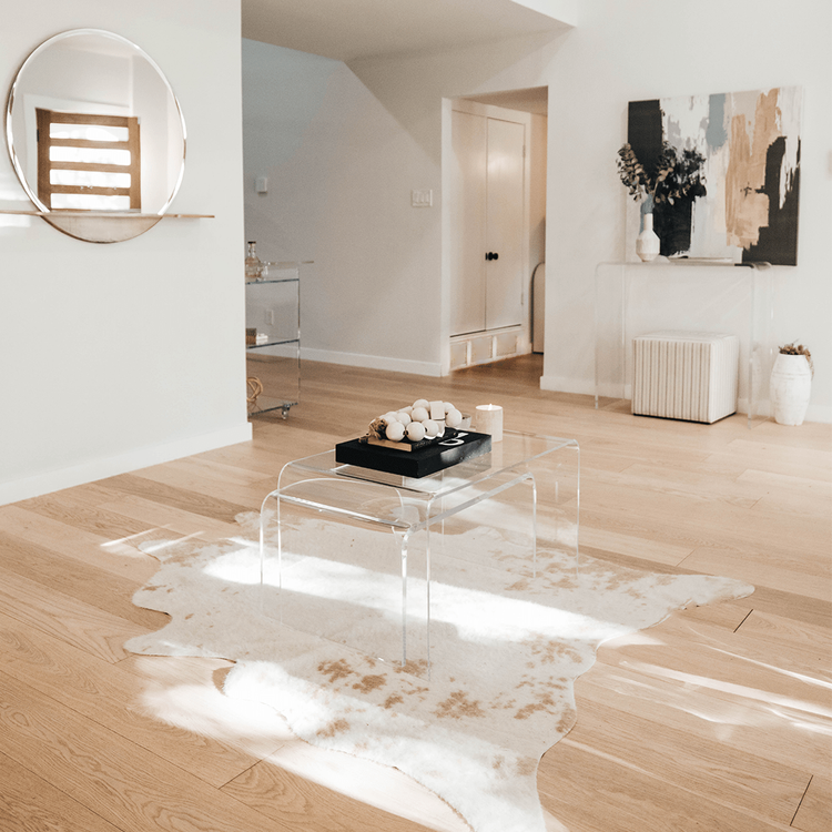 A clear acrylic lucite nesting coffee table set with candles, decor, and books on top placed on a light white and brown cowhide rug in a living room setting.