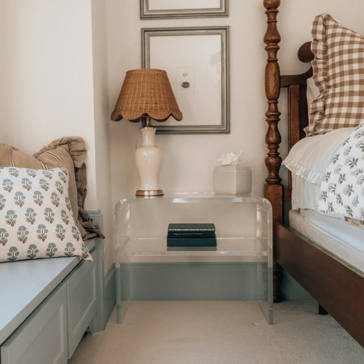 Clear acrylic side table with an interior shelf with decorative items and a lamp placed next to a bed.