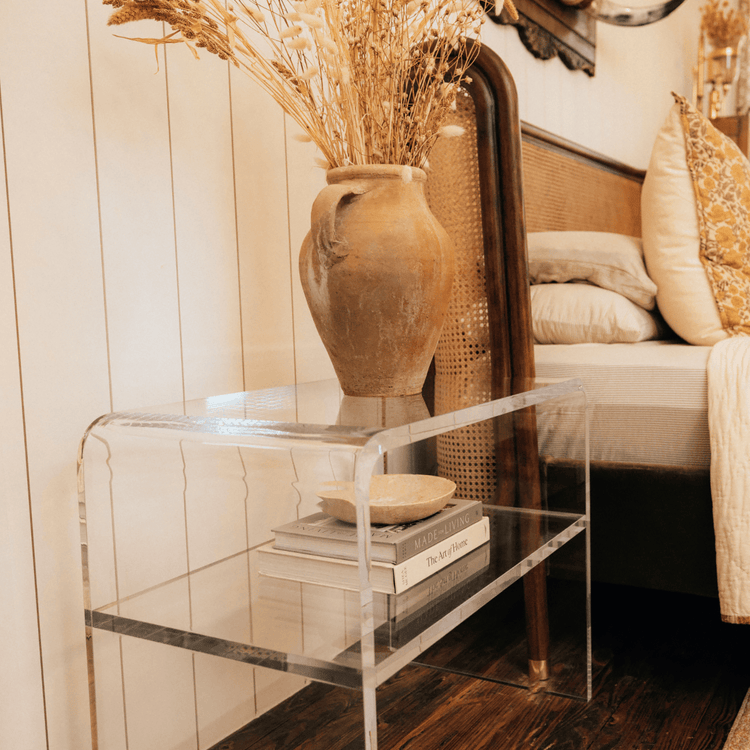 Clear acrylic side table with an interior shelf with decorative items placed next to a bed.