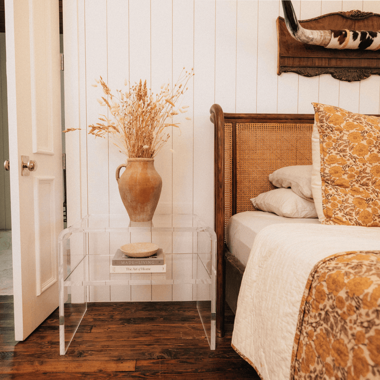 Clear acrylic side table with an interior shelf with decorative items placed next to a bed.