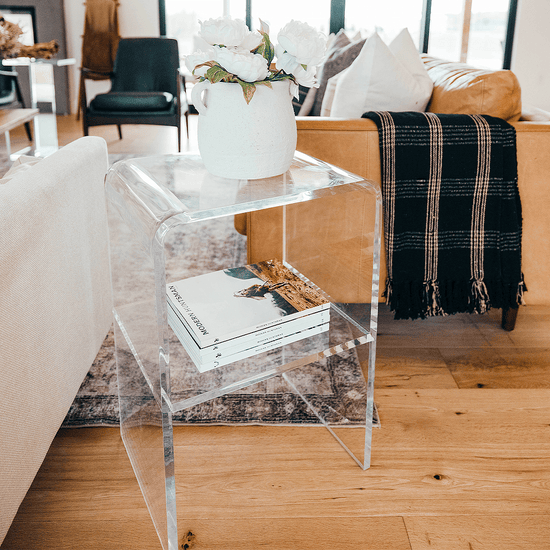 A clear acrylic lucite side table with waterfall edges and an interior shelf holding a vase of flowers and books placed between 2 couches in a living room setting.