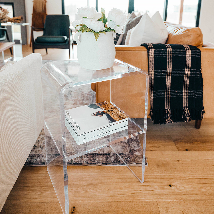 A clear acrylic lucite side table with waterfall edges and an interior shelf holding a vase of flowers and books placed between 2 couches in a living room setting.