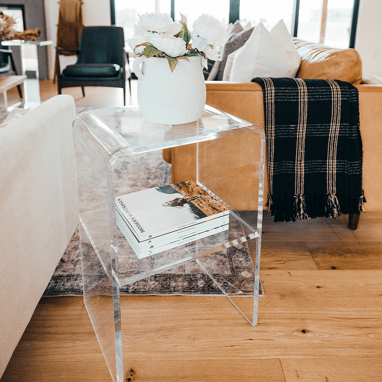 A clear acrylic lucite side table with waterfall edges and an interior shelf holding a vase of flowers and books placed between 2 couches in a living room setting.