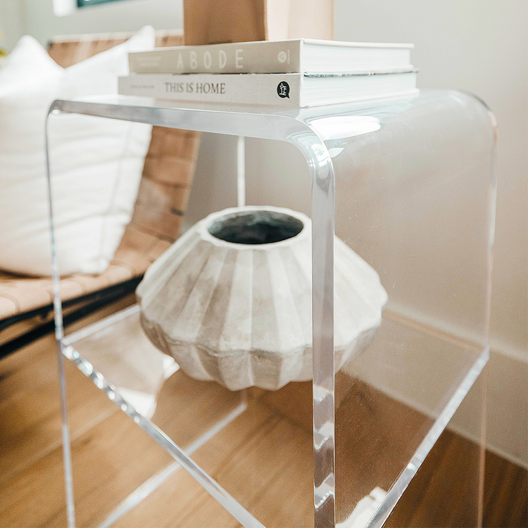 A clear acrylic lucite side table with waterfall edges and an interior shelf holding a vase and books placed beside a chair.