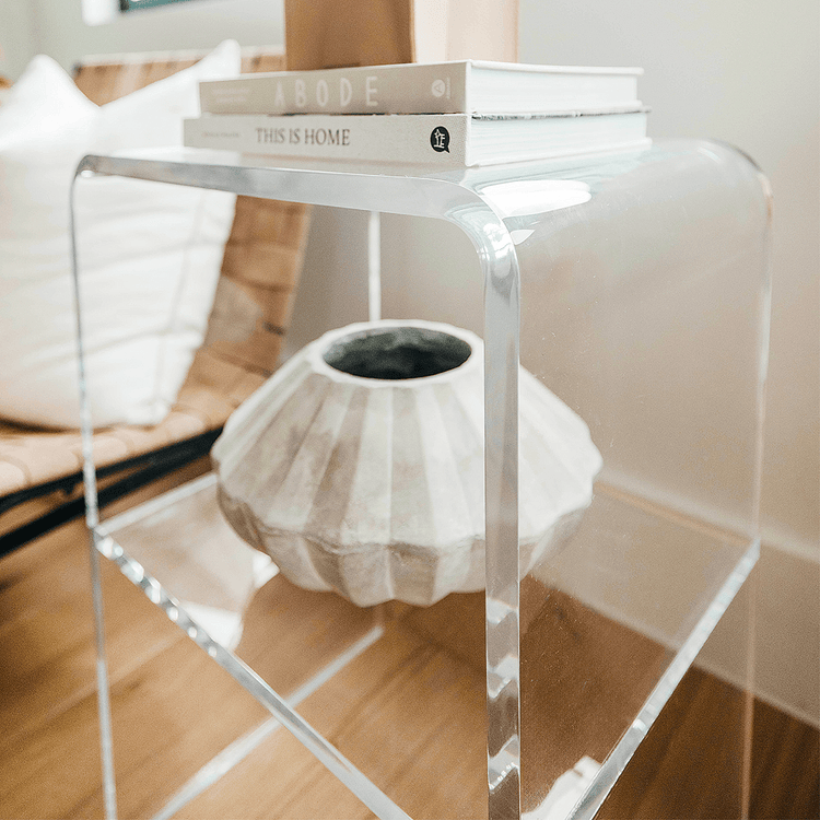 A clear acrylic lucite side table with waterfall edges and an interior shelf holding a vase and books placed beside a chair.
