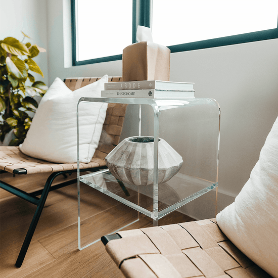 A clear acrylic lucite side table with waterfall edges and an interior shelf holding a vase and books placed beside a chair.