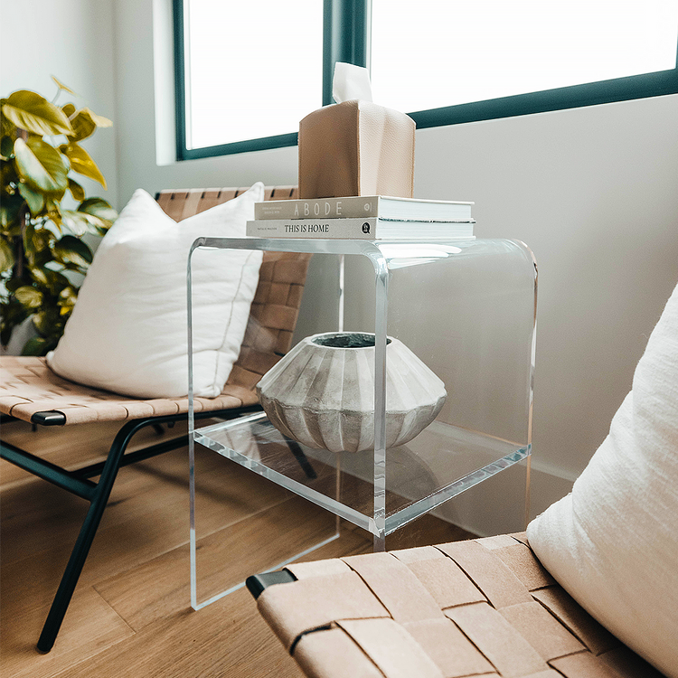 A clear acrylic lucite side table with waterfall edges and an interior shelf holding a vase and books placed beside a chair.