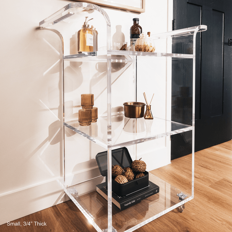 A clear acrylic bar cart featuring 3 shelves, wheels, and clear acrylic handles decorated with bar essentials, candles, and other decor items placed against a living room wall.