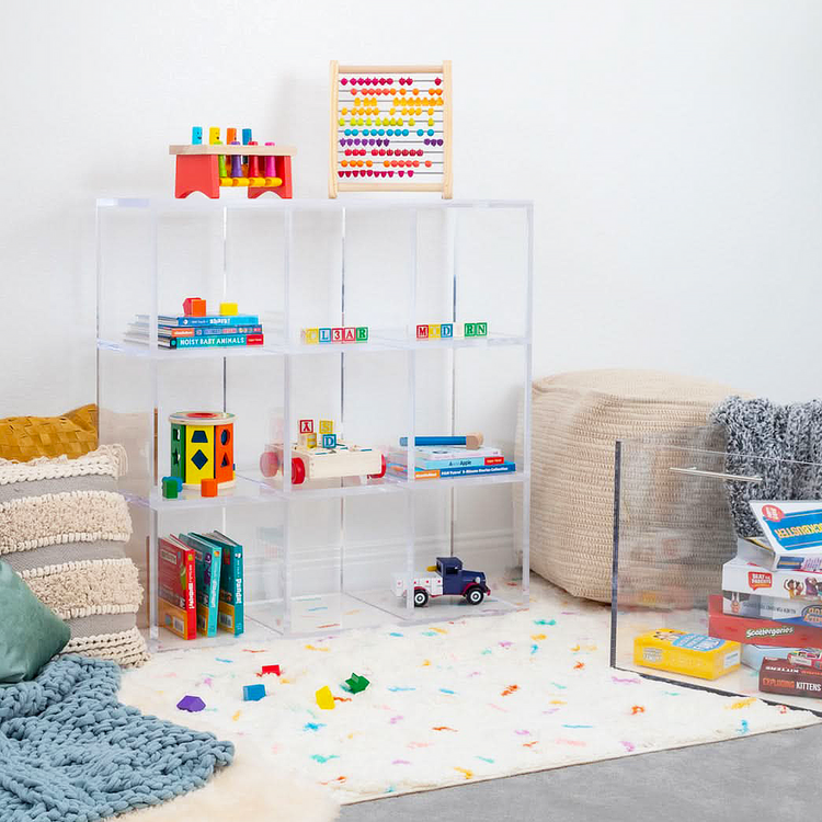 Clear acrylic 9 cube storage unit displaying children's toys, games, and books in a playroom furnished with pillows and an area rug. The unit features 3 rows of 3 cubes.