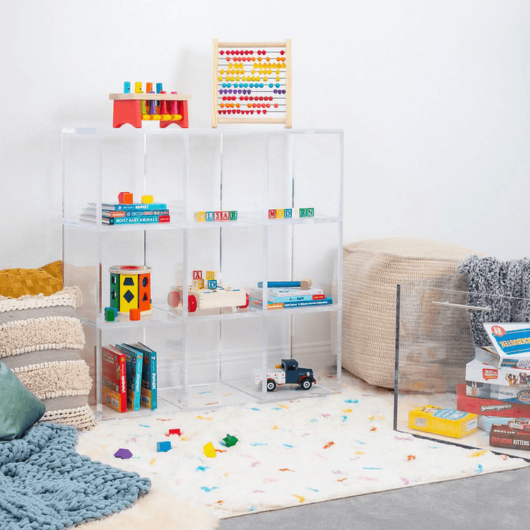 Clear acrylic 9 cube storage unit displaying children's toys, games, and books in a playroom furnished with pillows and an area rug. The unit features 3 rows of 3 cubes.