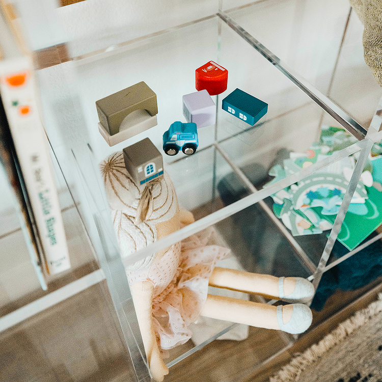 Closeup of a clear acrylic lucite 9 cube storage unit displaying children's toys, blankets, and books in a child's bedroom. The unit features 3 rows of 3 cubes.