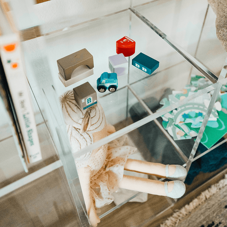 Closeup of a clear acrylic lucite 9 cube storage unit displaying children's toys, blankets, and books in a child's bedroom. The unit features 3 rows of 3 cubes.