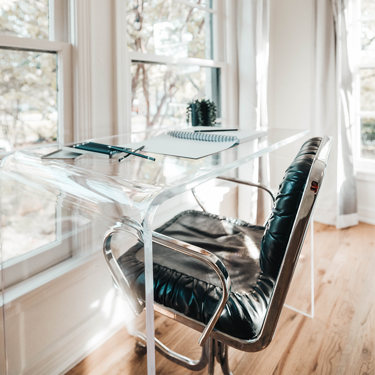 A clear acrylic waterfall edge vanity or desk displaying notebooks, pens, and a plant placed in front of a large bedroom picture window with a chair.