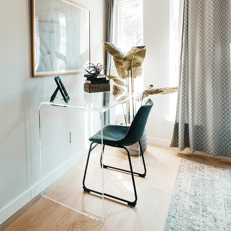 Clear acrylic waterfall work desk or computer desk decorated with photo frame and books next to a wall and windows.