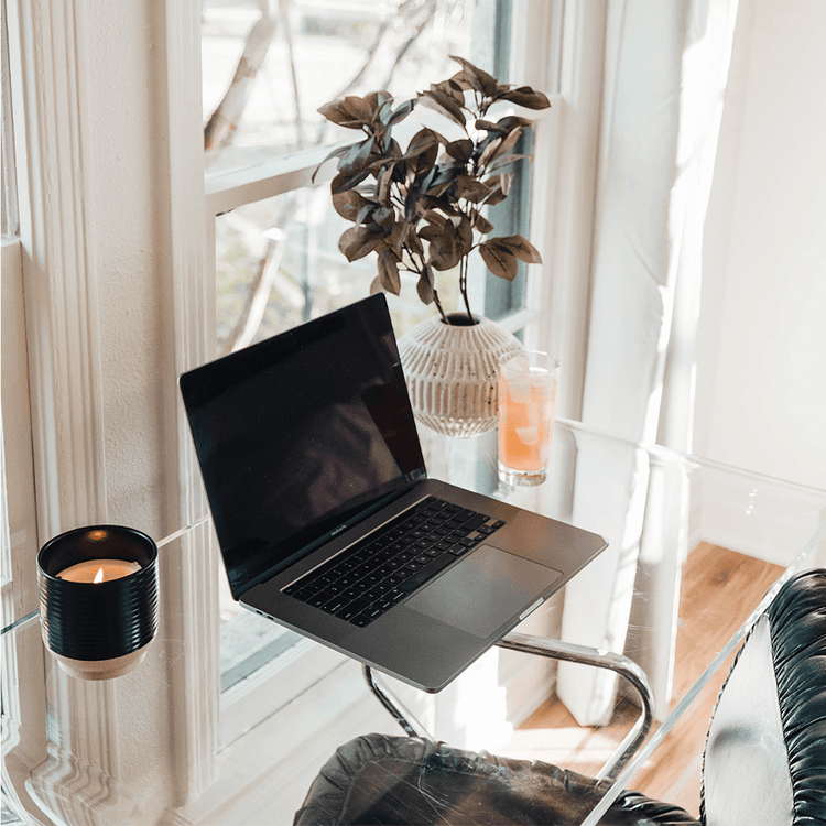 A clear acrylic waterfall edge vanity or desk displaying a laptop, a coffee cup, a plant, and a glass of juice placed in front of a large bedroom picture window with a chair.