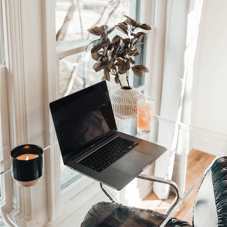 A clear acrylic waterfall desk in front of a window decorated with a candle, plant, glass of juice, and a laptop.