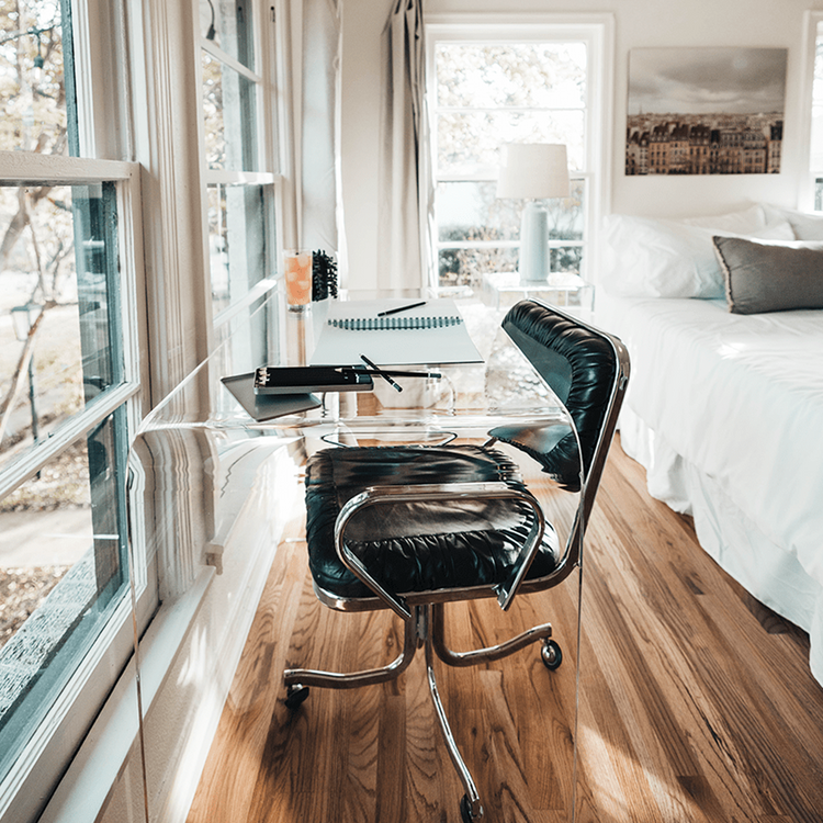 A clear acrylic waterfall desk decorated with various office supplies. a glass of juice, and chair pushed underneath facing windows.