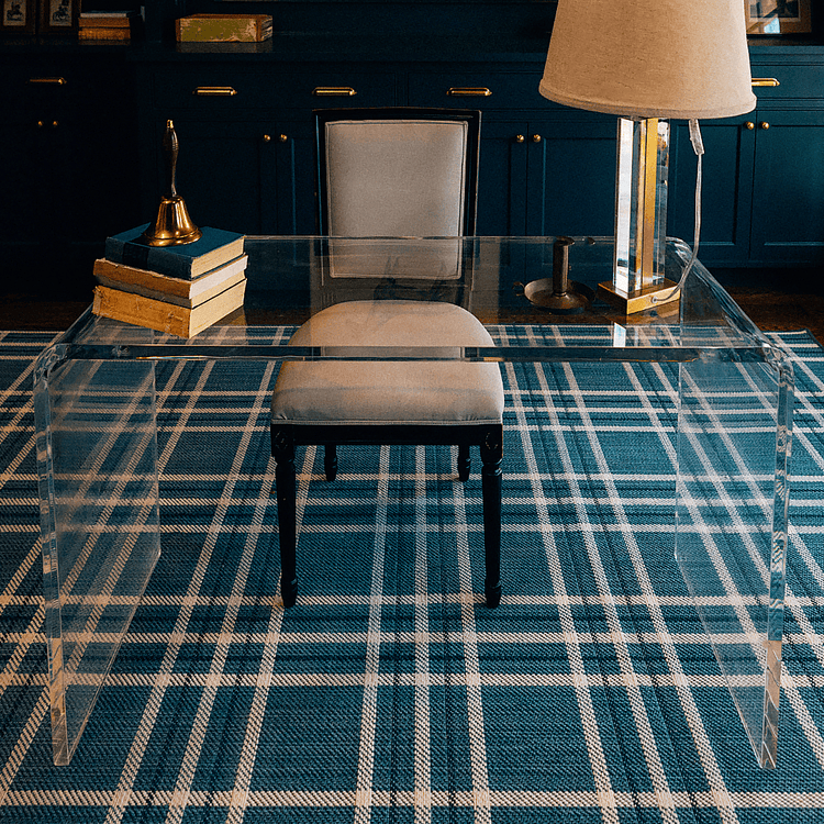 A clear acrylic waterfall edge desk placed on a plaid rug in a modern office setting on with a chair pushed underneath.