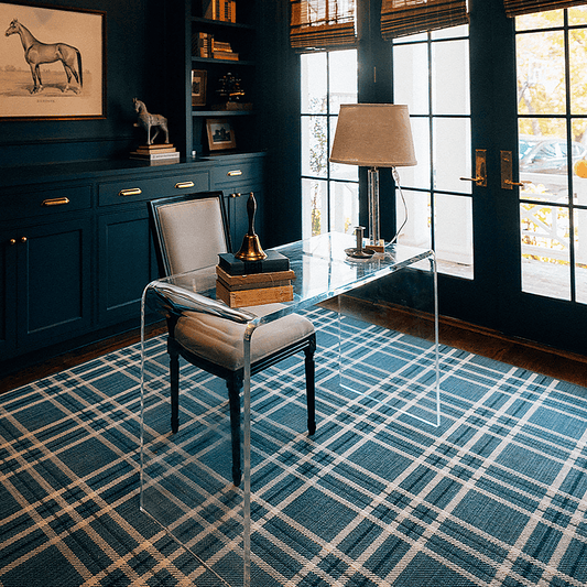 A clear acrylic waterfall edge desk placed on a plaid rug in a modern office setting on with a chair pushed underneath.