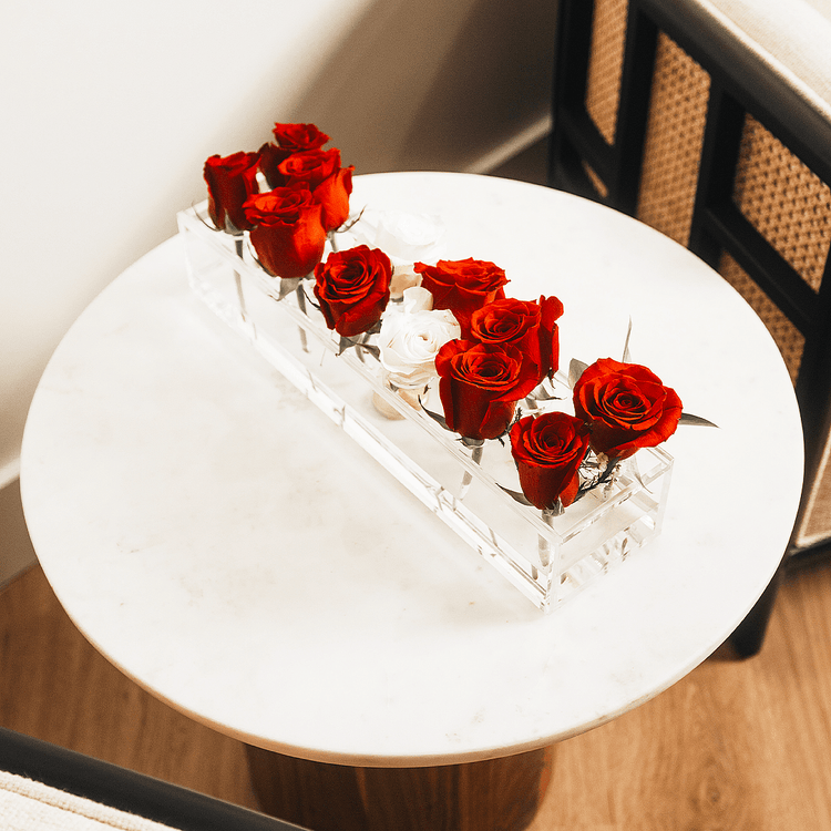 A clear acrylic horizonal vase holding 12 red and white roses on an end table next to a chair.