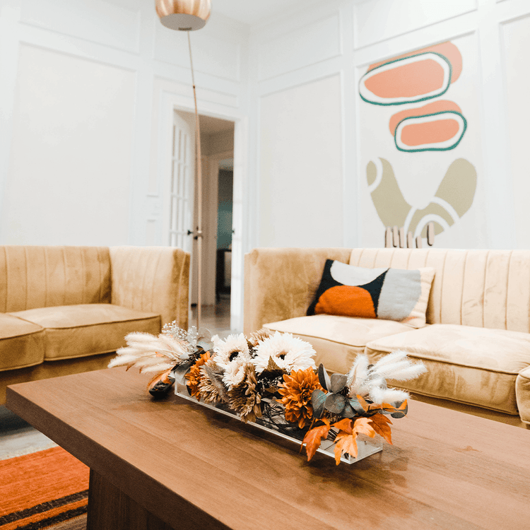 A decorative fall arrangement in a clear acrylic horizontal vase on a coffee table in a living room with beige couches and abstract wall art.