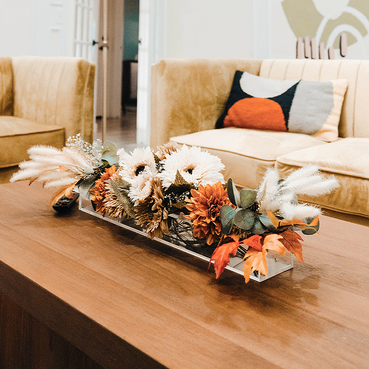 A decorative fall arrangement in a clear acrylic horizontal vase on a coffee table in a living room with beige couches and abstract wall art.