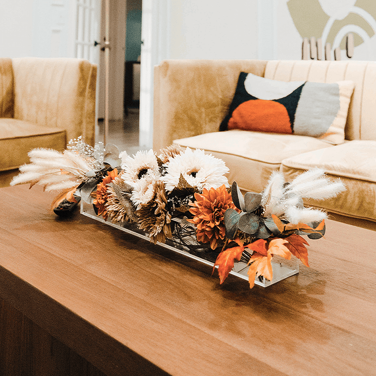 A decorative fall arrangement in a clear acrylic horizontal vase on a coffee table in a living room with beige couches and abstract wall art.
