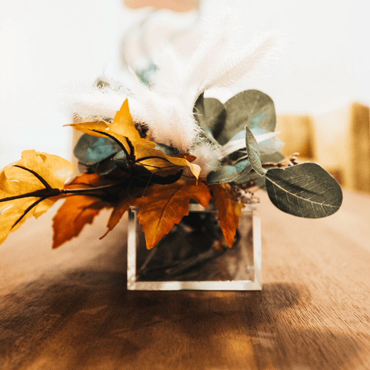 A closeup of a decorative fall arrangement in a clear acrylic horizontal vase on a coffee table in a living room.