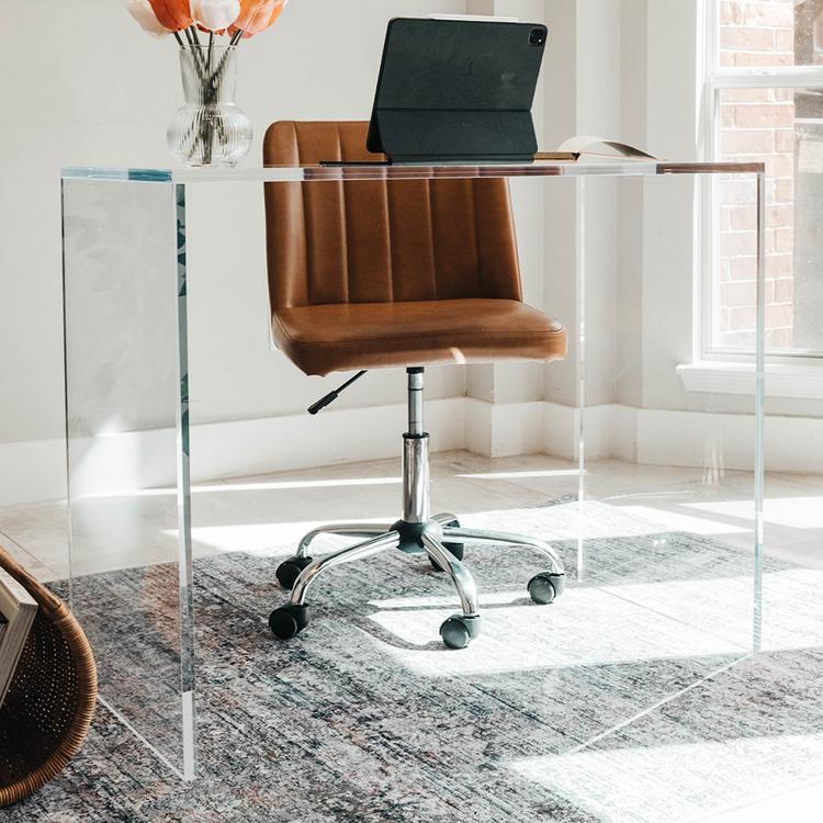An acrylic lucite slab desk with a computer on top and a brown leather chair in front, placed in an office setting.