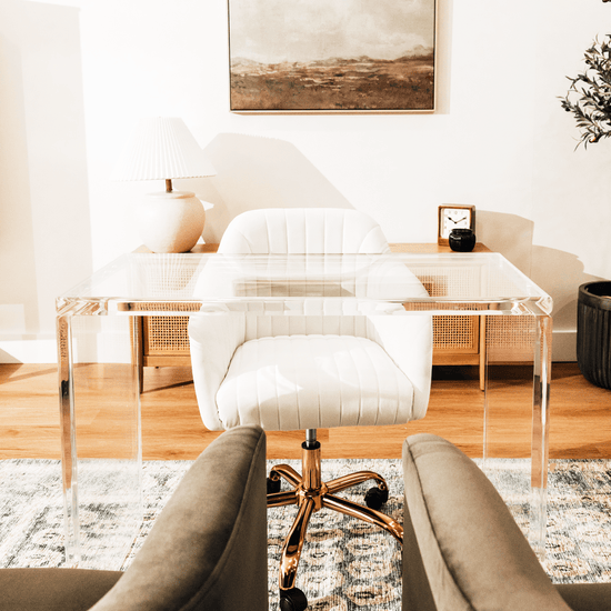 An acrylic lucite slab desk with a white chair in front, placed in an office setting with a painting on the wall and a tree in the background.