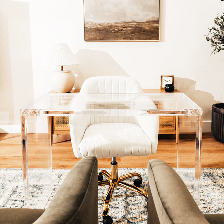 An acrylic lucite slab desk with a white chair in front, placed in an office setting with a painting on the wall and a tree in the background.
