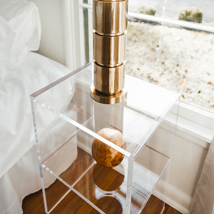 Closeup looking down through a clear acrylic slab nightstand with a lamp placed on top and 1 interior shelf in front of a window next to a bed.