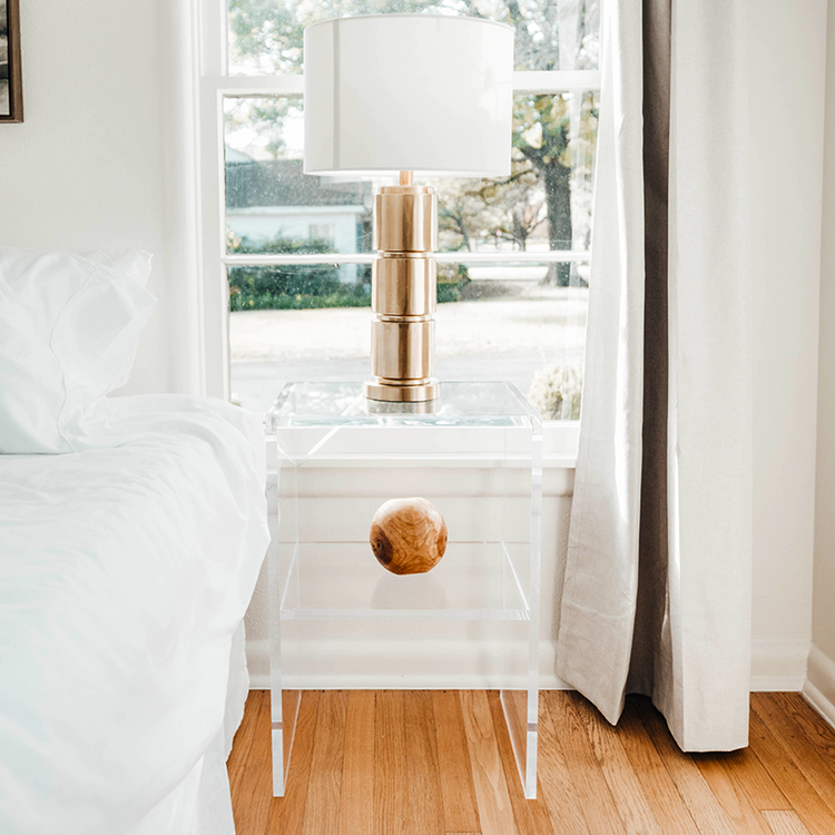 Clear acrylic lucite nightstand with gold lamp and decorative object next to a bed in a bedroom.