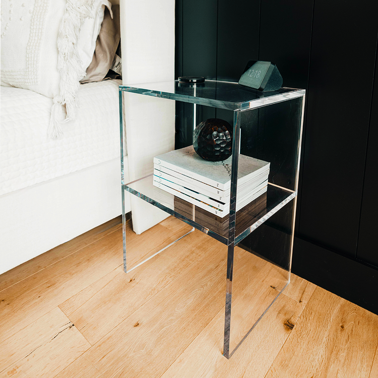Clear acrylic lucite nightstand with 1 interior shelf holding an alarm clock, books, and decorative items in a bedroom setting.
